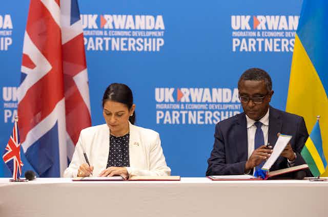 A woman and a man sign documents behind a press conference table. Behind them stand the British and the Rwandan flags.