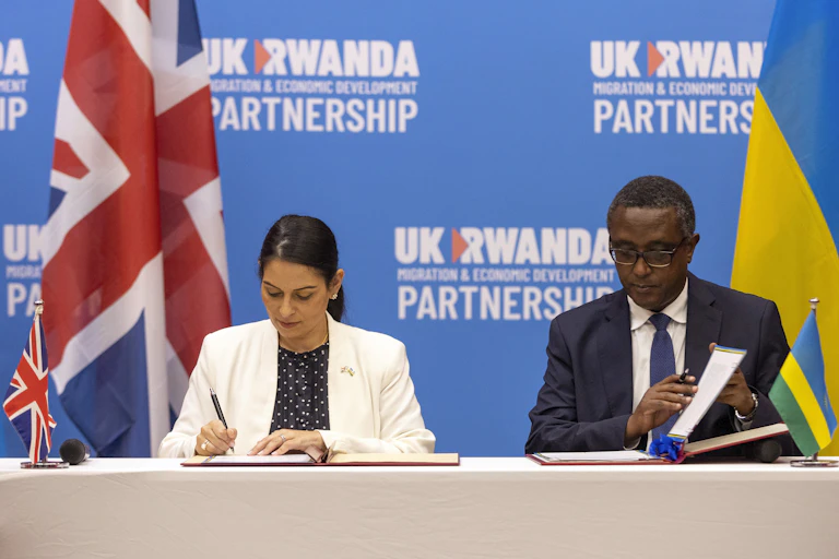 A woman and a man sign documents behind a press conference table. Behind them stand the British and the Rwandan flags.
