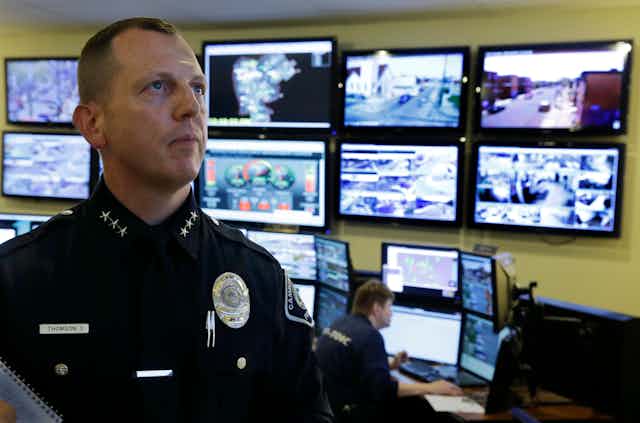 a man in a police uniform in the foreground looks up as a man seated in the background watch is a video monitor and the wall in the background is covered in video monitors