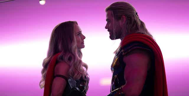 Two superheroes seen lovingly gazing at each other in front of a purple background.