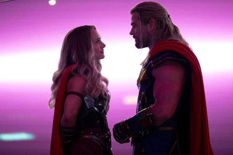 Two superheroes seen lovingly gazing at each other in front of a purple background.