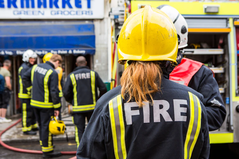 A female firefighter with a ponytail viewed from behind, wearing a yellow helmet and uniform that says FIRE on the back