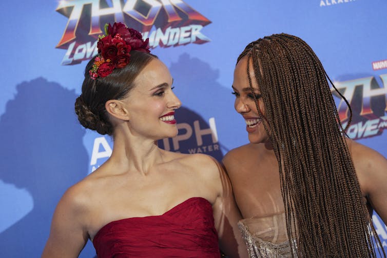 Two women in fancy dresses seen against a blue backdrop smiling at each other.