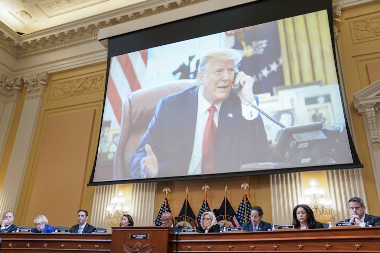 A middle-aged man wearing a navy blue suit, white shirt and red tie is seen on a large screen talking on a telephone.