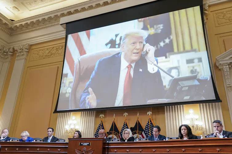 A middle-aged man wearing a navy blue suit, white shirt and red tie is seen on a large screen talking on a telephone.