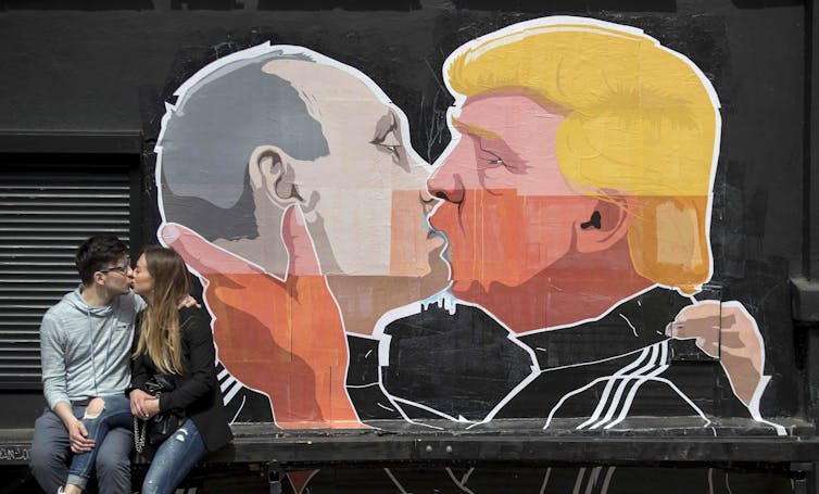 A couple kisses in front of a graffiti mural of two men kissing, one with gold hair.