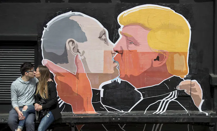 A couple kisses in front of a graffiti mural of two men kissing, one with gold hair.