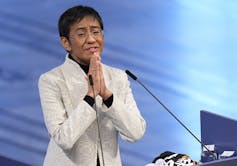 A woman with short dark hair in a white jacket clasps her hands together as she stands in front of a microphone.