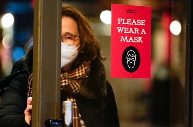 A woman in a face mask opening a door. There is a sign on the door that says 'Please wear a mask'