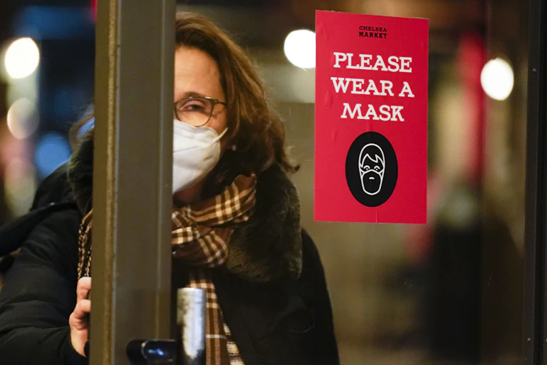A woman in a face mask opening a door. There is a sign on the door that says 'Please wear a mask'