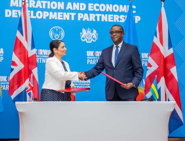 With the British and Rwandan flags in the background, a Black man and a white woman shake hands after signing migration documents 
