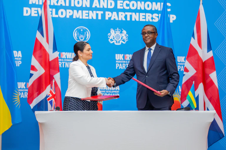 With the British and Rwandan flags in the background, a Black man and a white woman shake hands after signing migration documents