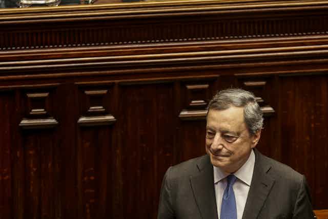 Italian Premier Mario Draghi smiles while wearing a grey suit, standing in front of a wood panel.