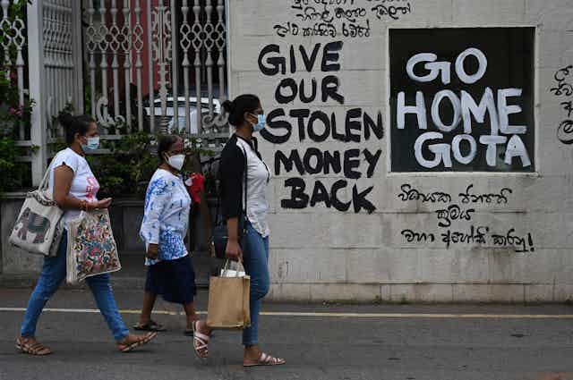 Three women wearing masks walk past a wall with graffitied slogans that read 'Give Our Stolen Money Back' and 'Go Home Gota'