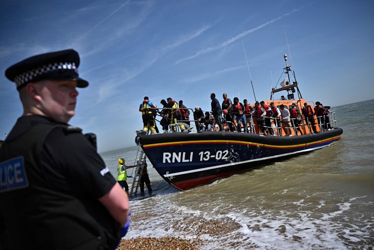 a police officer stands on the beach and watches people get off of a lifeboat.