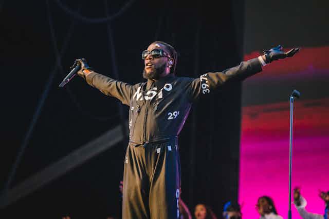 A man with dreadlocks, shades and a beard wears a black boiler suit, his arms outstretched with stage lights behind him.