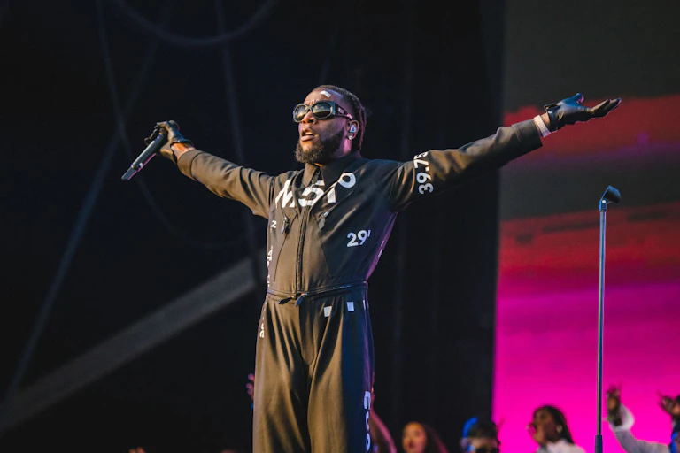 A man with dreadlocks, shades and a beard wears a black boiler suit, his arms outstretched with stage lights behind him.
