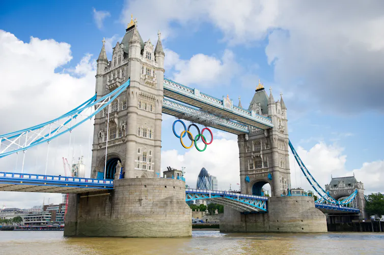Tower Bridge seen against a blue and cloudy sky, with the Olympic rings suspended from the main structure.
