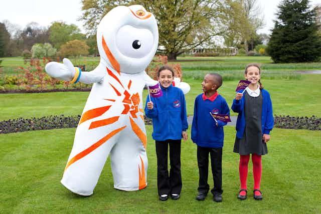 A white and orange costumed character in a garden with school children in uniform holding tiny flags.