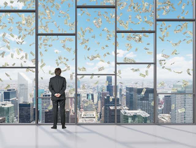 Suited man watches through high window as bank notes fall through the sky.