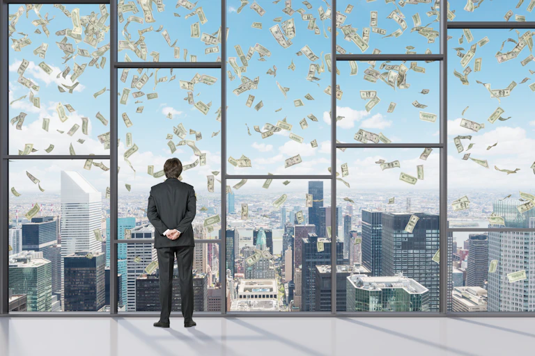 Suited man watches through high window as bank notes fall through the sky.