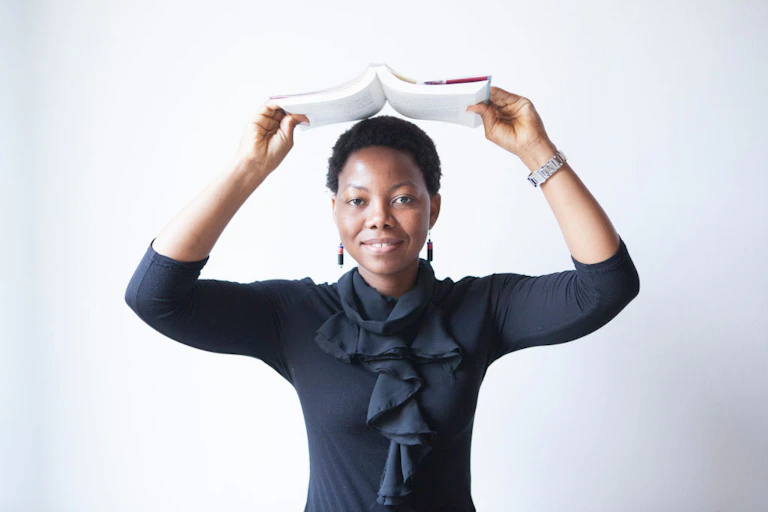A young woman in a black blouse holds a book open above her head, a small smile on her face.