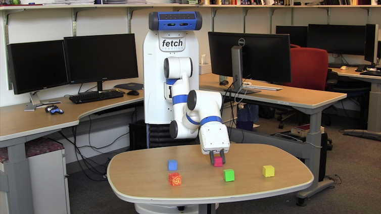 a robot with a single arm grasps one of five colored blocks on a small table