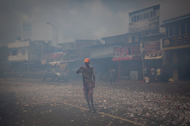 A man in an orange cap stands in a smoky street surrounded by strewn bits of paper.