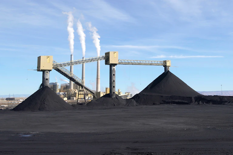 A power plant with three active smokestacks and three large piles of coal in front of it.