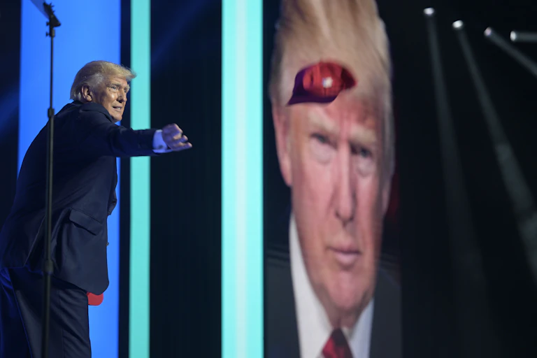 A man with blond-grey hair in a dark suit tosses a red ball cap. A large photo of his face is in the background.