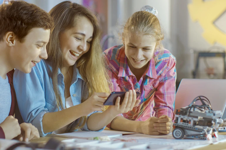 Dos chicos y una chica utilizan un teléfono móvil en un aula.