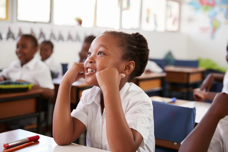 A young girl in a white shirt, sitting at a desk, with other children in the background in a classroom