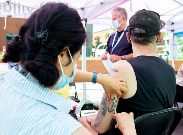 A person injects a vaccine into the back of the arm of a a man wearing a black tank top and a black baseball cap.
