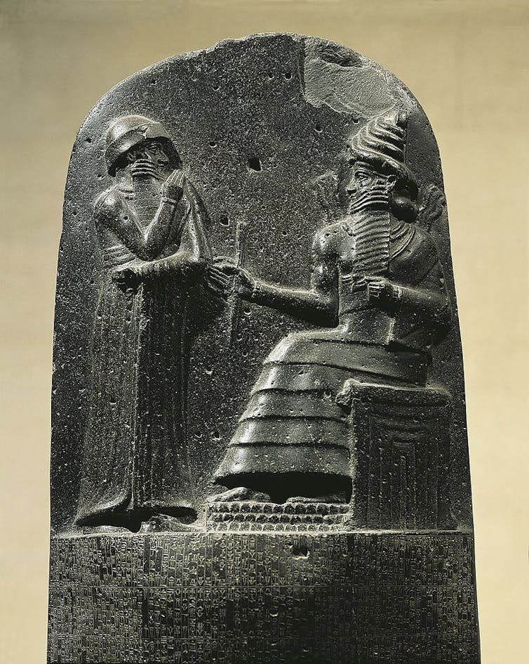 A stone relief shows two men with long beards: one standing, with a hand to his mouth, the other seated and holding a staff.