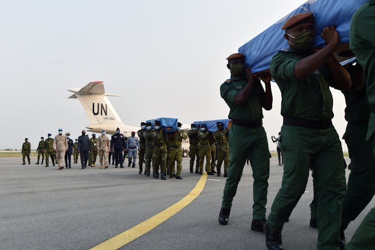 Soldiers are seen carrying coffins draped in blue flags, in front of a white UN plane.