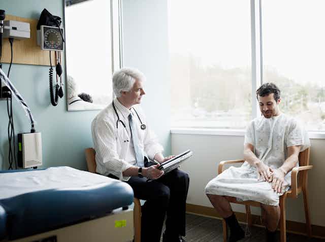 An older, white-haired, white doctor with a stethoscope hanging around his neck sits near a very young, dark-haired white man wearing as hospital gown and looking at his hands as he talks, as if he's a little embarrassed, a little nervous and wondering why he feels a draft.