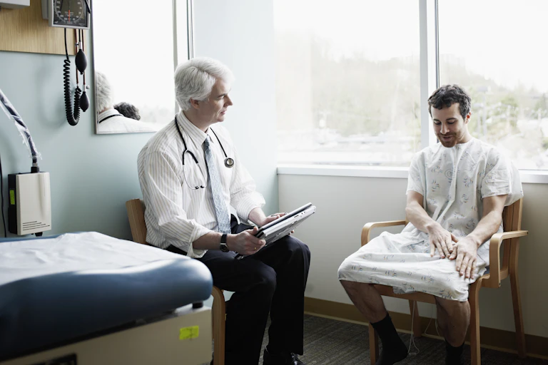 An older, white-haired, white doctor with a stethoscope hanging around his neck sits near a very young, dark-haired white man wearing as hospital gown and looking at his hands as he talks, as if he's a little embarrassed, a little nervous and wondering why he feels a draft.