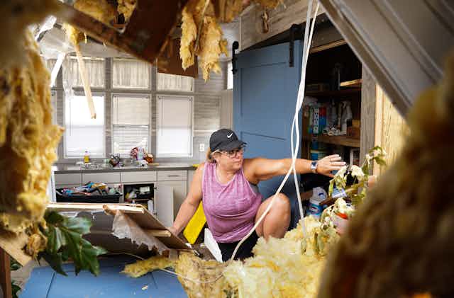 A woman climbs through a hurricane-damaged kitchen. A hole is evident in the side and roof of the house, with insulation hanging out.