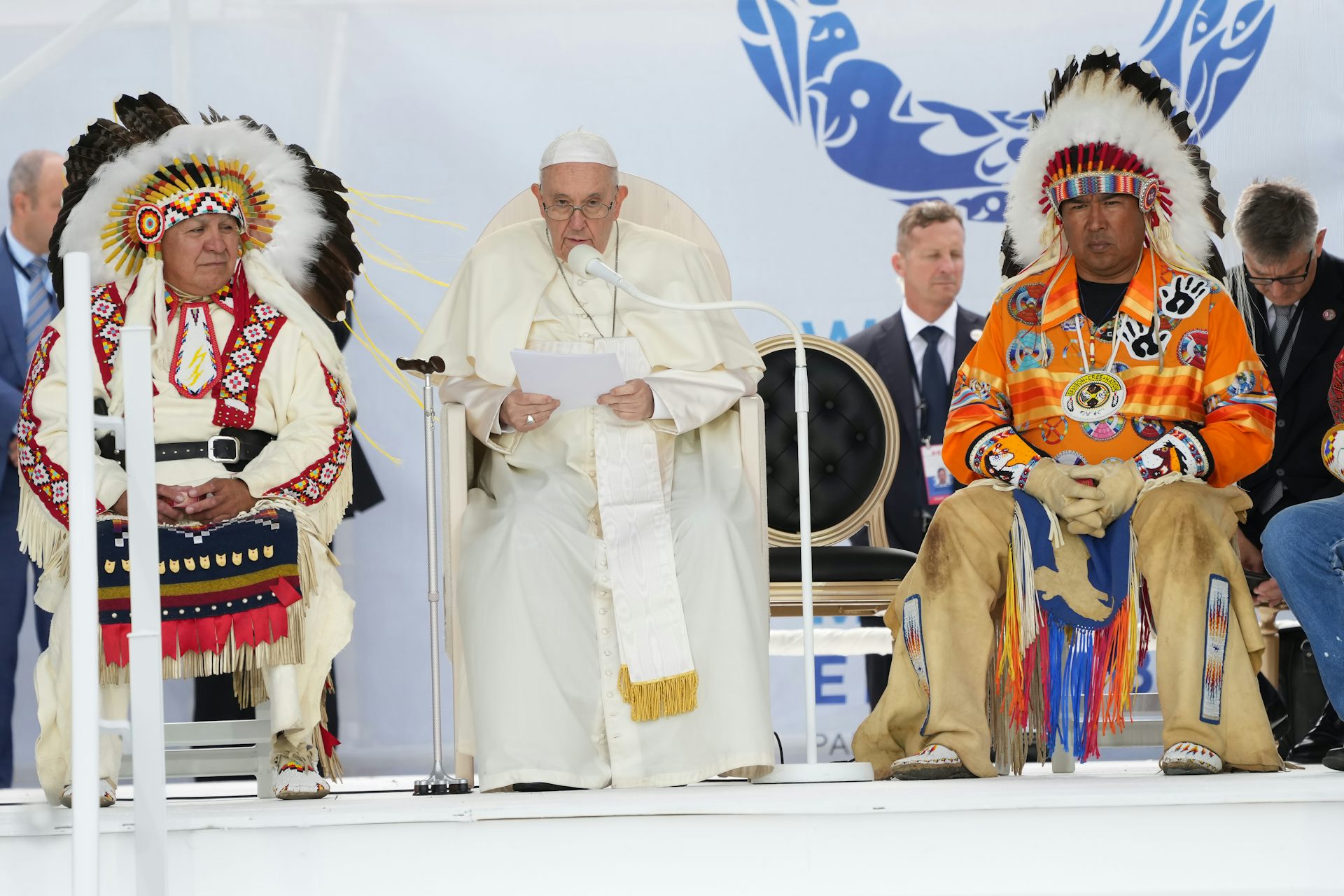 The Pope, in a white skullcap and  robes, is seen seated, flanked by two men in regalia including headdresses.
