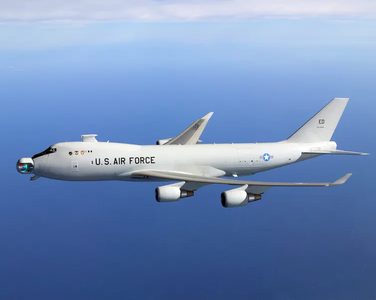 A large four-engine jet aircraft painted light gray and a large iridescent object attached to its nose
