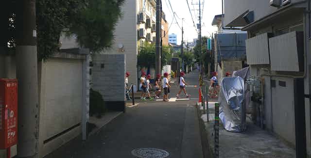 A group of primary school children walk along a quiet, narrow street in Nakano, Japan.