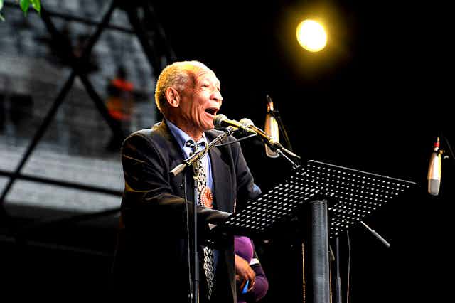 An elderly man in a suit and tie speaks into a microphone on a stage. He appears to speak effusively, smiling as he does.