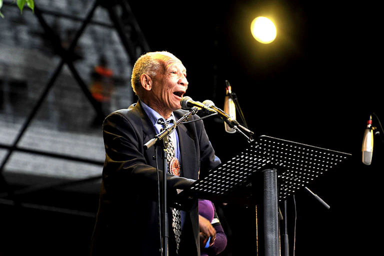 An elderly man in a suit and tie speaks into a microphone on a stage. He appears to speak effusively, smiling as he does.