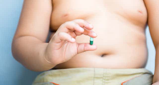 Una persona obesa con el torso a la vista muestra una píldora verde en la mano.