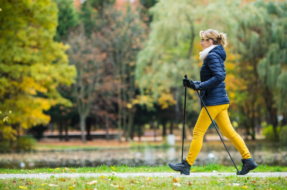A woman wearing a winter jacket uses Nordic walking poles during her walk through a park.