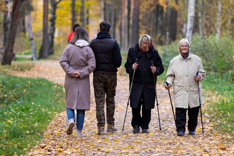 Two elderly ladies wearing winter coats stroll down a woodland path using walking poles.