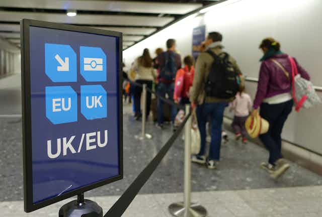 A UK/EU passport lane sign at an airport, with families walking by