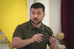 A dark-haired man with a beard wearing a dark green short-sleeved shirt speaks into a microphone.