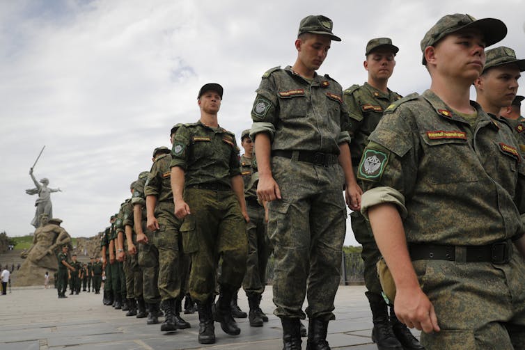 A row of Russian soldiers in green camouflaged army garb parade with a monument behind them.