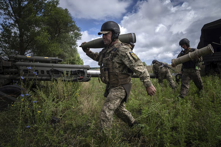 A soldier carries an artillery shell on his shoulder in a rural area, with trees in the background.